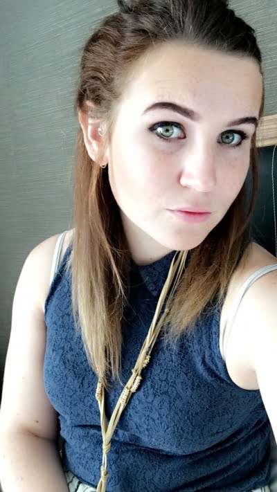 Young lady wearing a blue top, long necklace. She has long dark hair tied back.