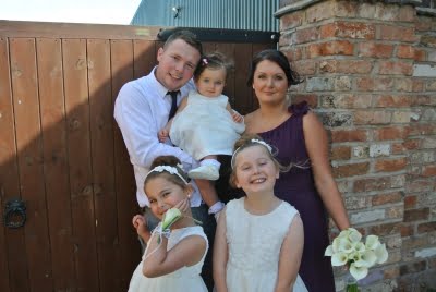 Family of five, man and lady with three daughters, the girls are wearing cream dresses, the Mum a dark dress and the Dad a white shirt and tie. They are in wedding attire.