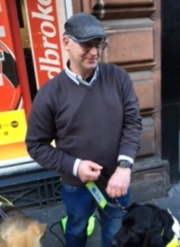 A man wearing a grey cap, are jumper and jeans. He is holding his Guidedogs lead.