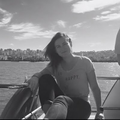 Black and white picture of a young lady sitting on a boat with the sea behind her. Her hair is blowing in the wind and she is smiling.