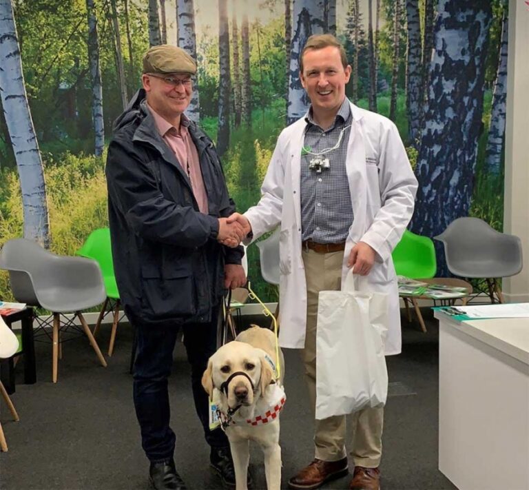 Two men and a golden guidedog. One man wearing a white doctor like gown and the other with a dark coat, brown cap and glasses, they ar shaking hands.