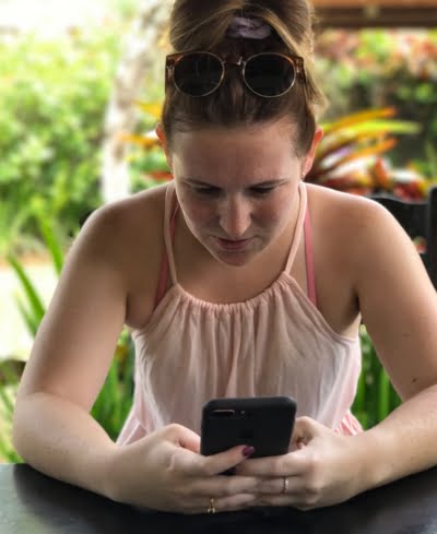 Young lady looking down at her iPhone, she is wearing her hair up and sunglasses on top of her head, she is wearing a pink top.