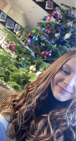 A smiling young lady with long hair sitting in front of a Christmas tree.