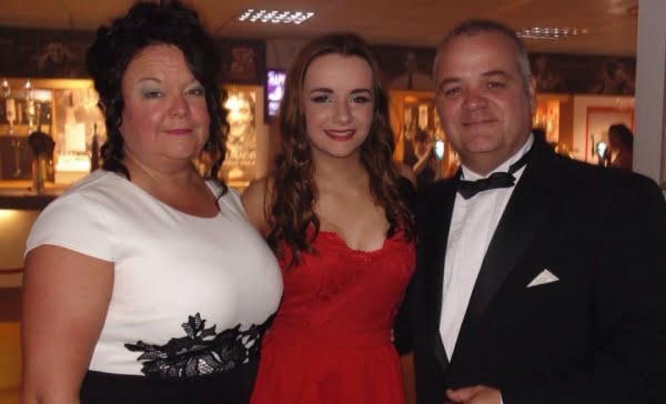 Jodie and her parents, Jodie is in the middle wearing a red dress, her mum a white dress with a little black detail and her dad a black suit jacket, white shirt and black bow tie