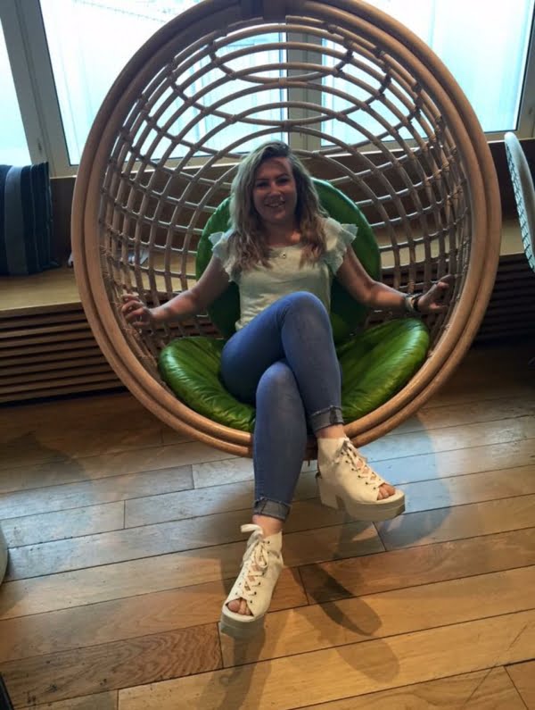 A young lady sitting in a hanging wicker swing, she has her legs crossed, wearing blue jeans, cream wedge shoes and light green top.