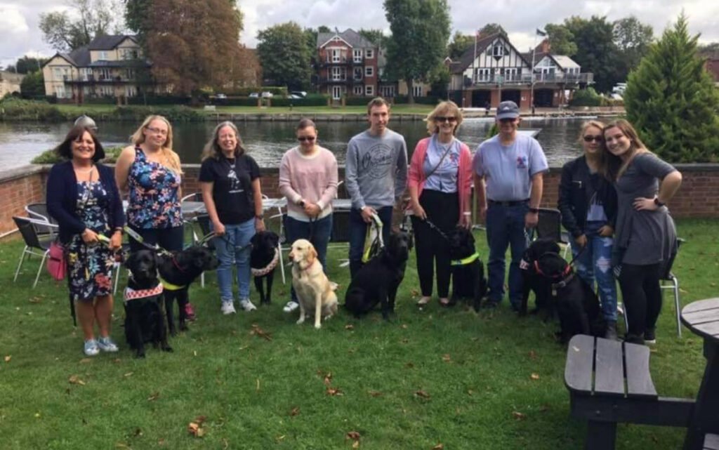 Group picture of seven ladies, two men, seven black Guidedogs and one golden coloured guidedog. They are standing on grass with river behind them.