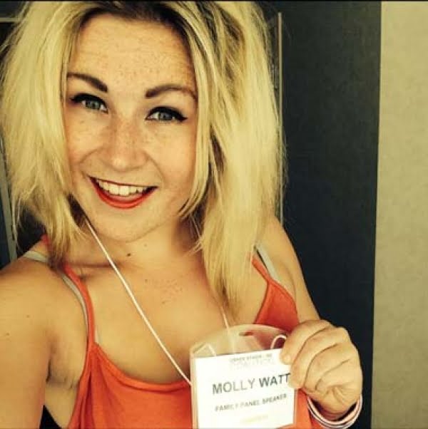 Molly smiling looking at the camera, wearing and orange vest top and holding her name tag which reads Molly Watt.