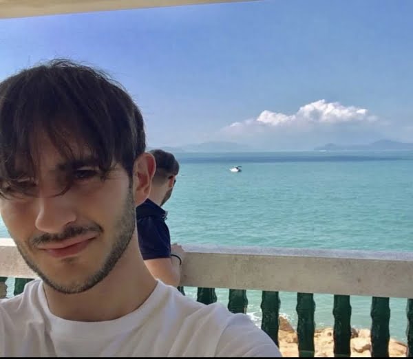 Dark haired man with some facial hair, behind him is the sea, blue sky with white clouds.