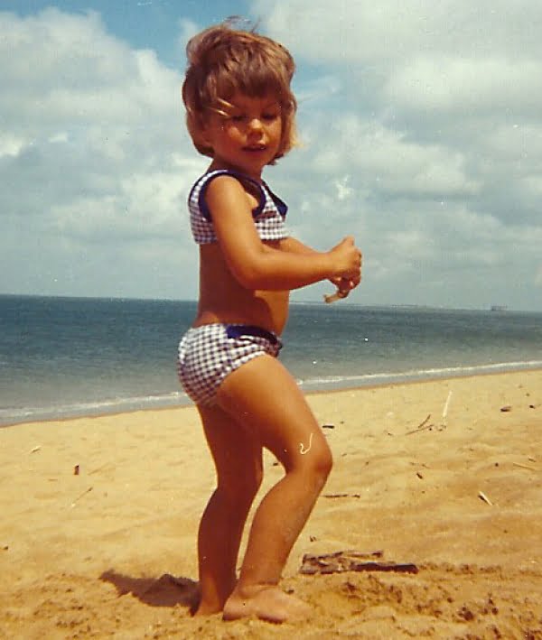A little brown haired girl wearing blue and white gingham bikini, she is on a sandy beach with the sea in the background.